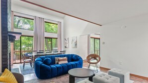 A modern living room with a blue sectional sofa, grey ottomans, and a round coffee table. There is a dining area with a table and chairs visible through large windows. - Hot Tub, Fire Pit, Game Room; 4BR Cozy Poconos! (East Stroudsburg)