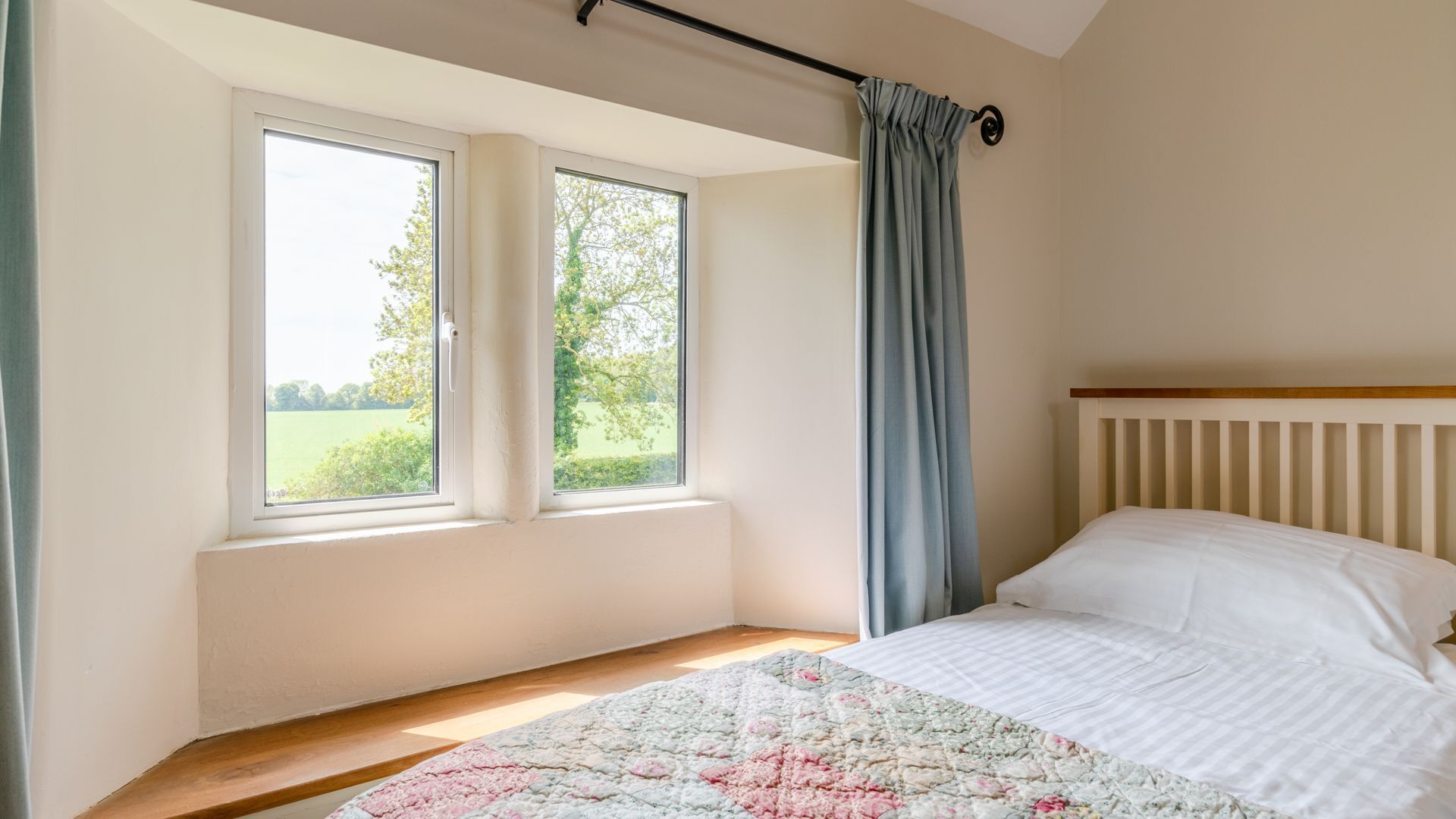 A bedroom with a bed, two windows, a view of greenery, and a curtain.