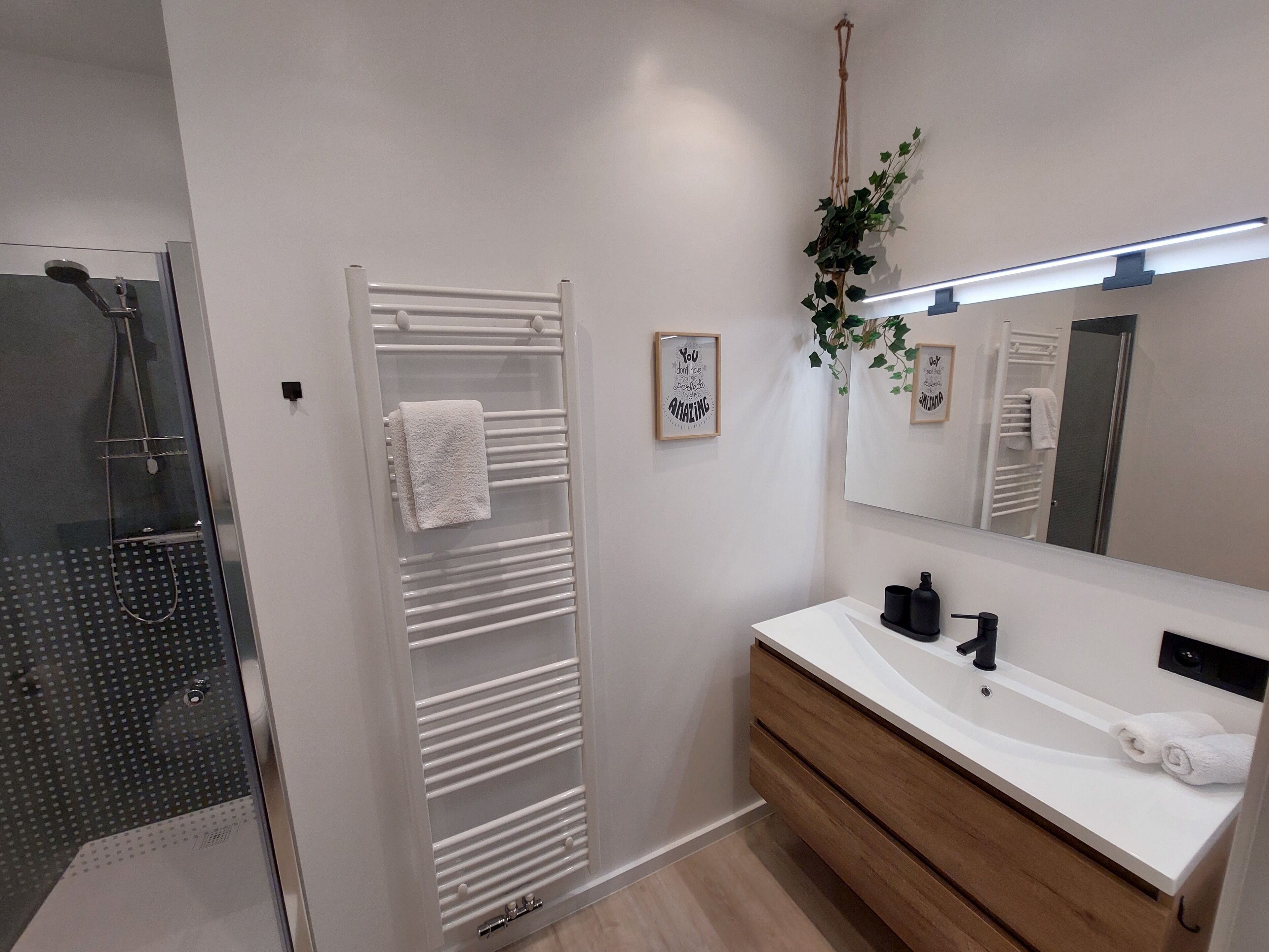 A modern bathroom with a glass shower enclosure, a white towel rack, a wooden vanity with a white countertop, and a large mirror.