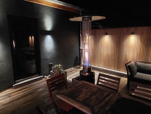 Um alpendre de madeira com pouca luz, com uma mesa e cadeiras, um lustre pendurado e vista para um quarto com porta de correr.