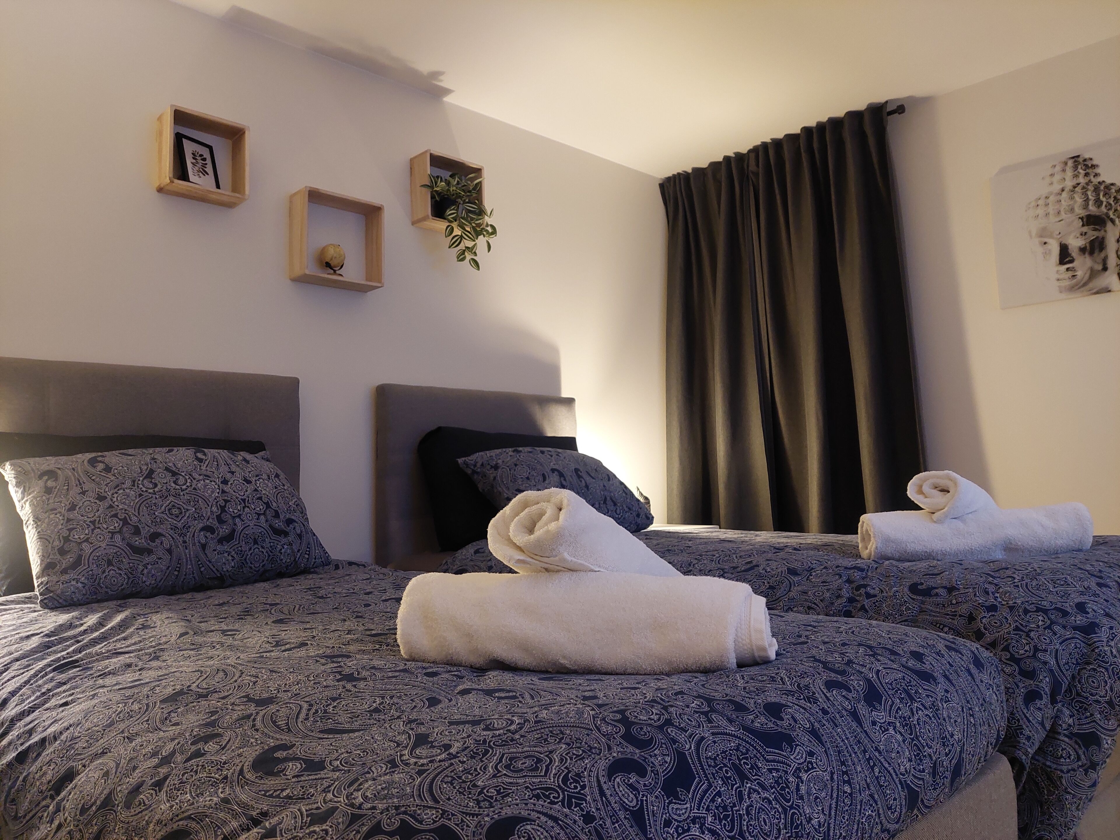 A hotel room with two beds, each with a patterned bedspread and matching pillows. There are three small wooden shelves with decorative items, a large window with dark curtains, and a wall-mounted artwork.