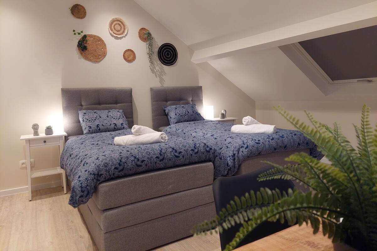A modern bedroom with a loft-style ceiling, two beds with matching blue patterned bedding, a grey upholstered bench, and decorative wall art.