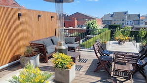 A wooden deck with outdoor furniture, including a sofa, chairs, and a table. There are flower planters and a decorative umbrella. - Viator (Tongeren)