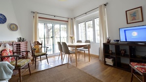 A spacious living room with a dining area, large windows, a television, and a bookshelf.