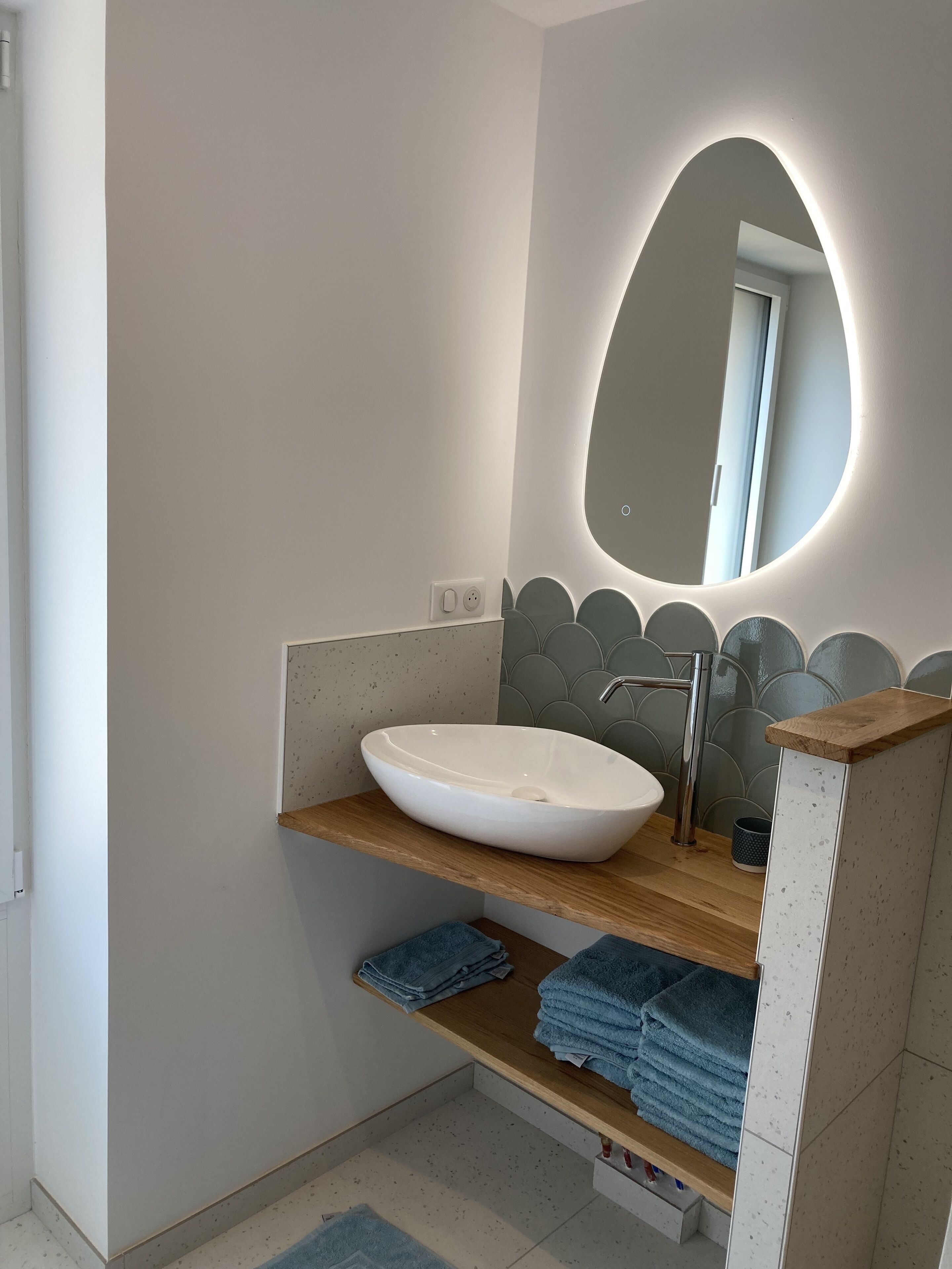 A modern bathroom with a white sink, a wooden countertop, a mirror with a scalloped edge, and a towel rack with folded towels.
