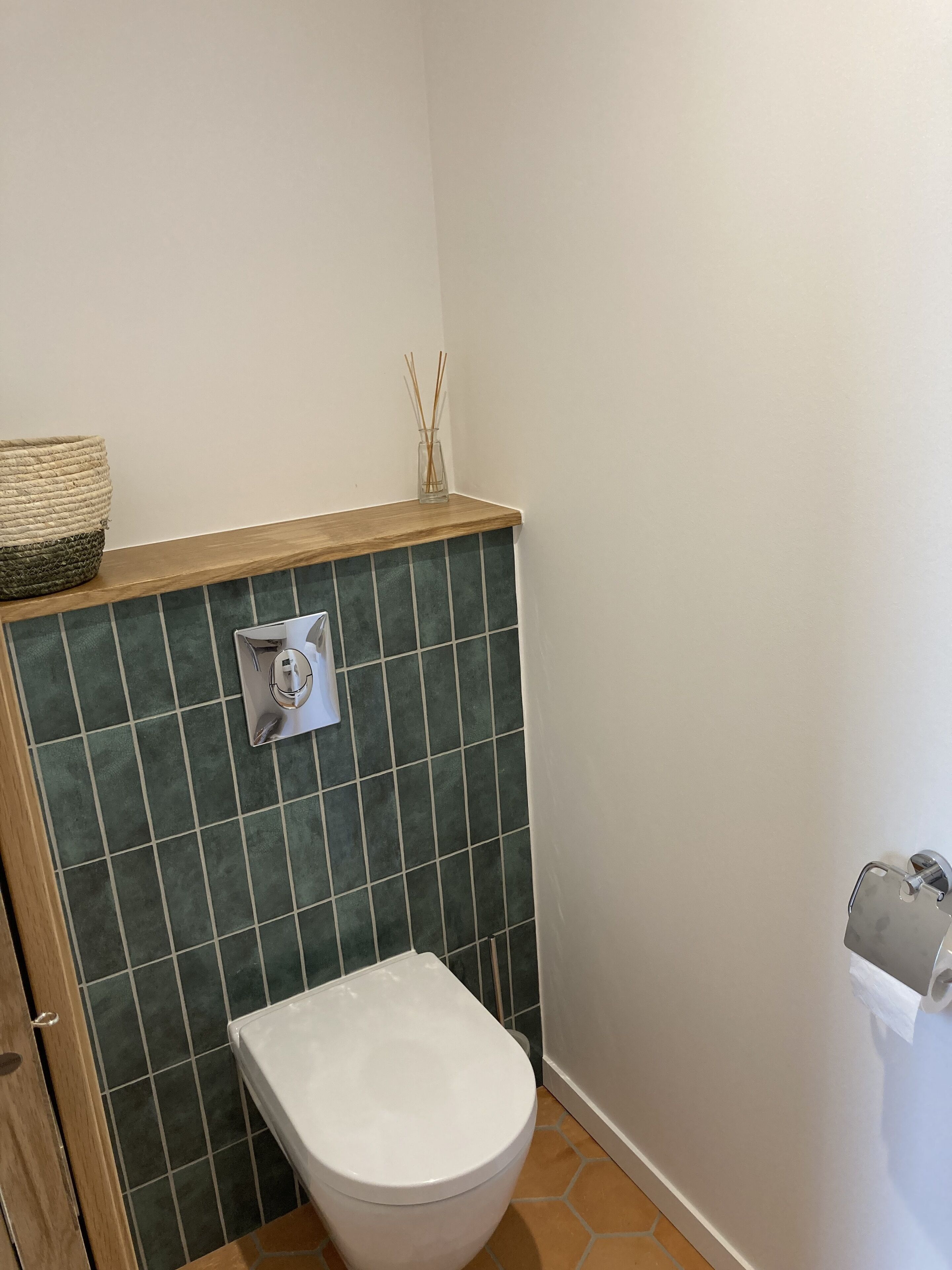 A bathroom with a white toilet, green tiled walls, a wooden shelf with a woven basket, and a toilet paper holder.
