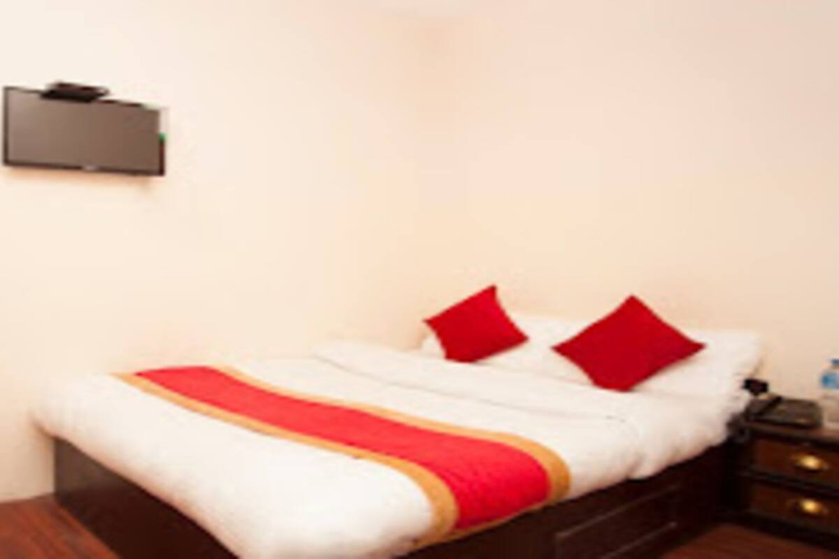 A hotel room with a bed, two red pillows, a white and red bedspread, a nightstand with water bottles, and a TV mounted on the wall.