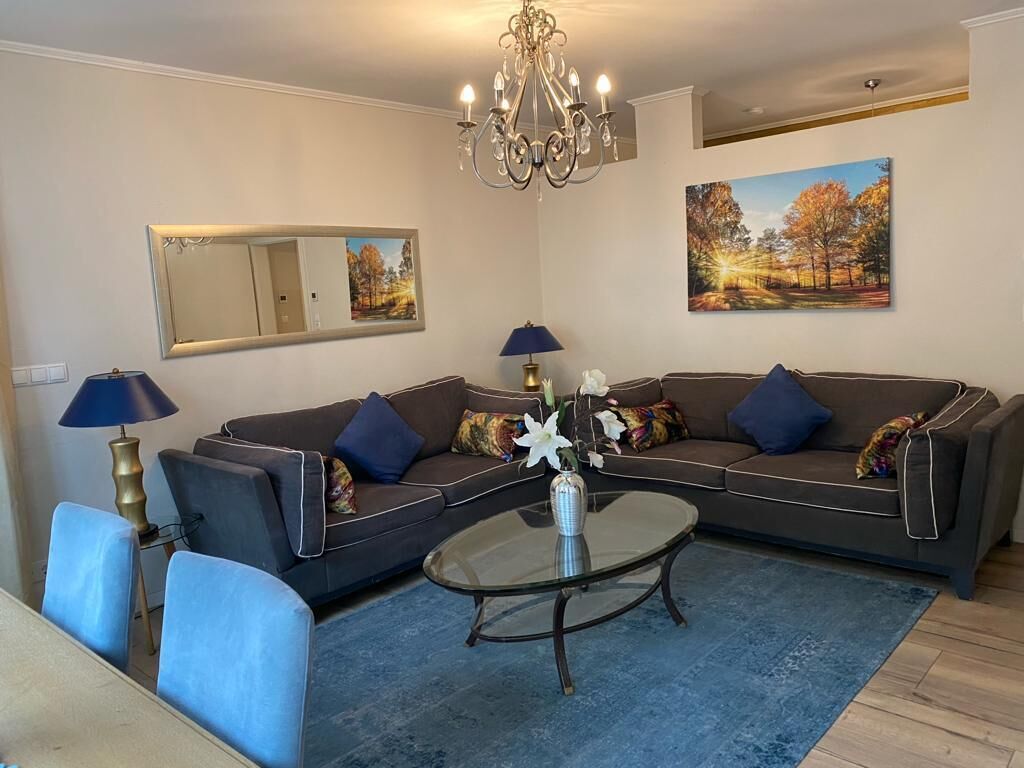 Ein Wohnzimmer mit einem Ecksofa, einem Glastisch, einer Deckenleuchte, einem Spiegel und zwei Bildern.