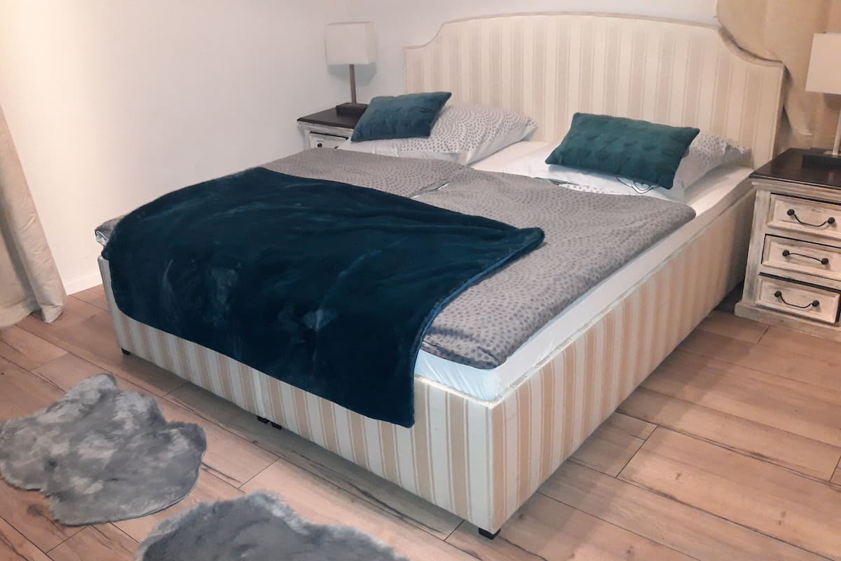 Uma cama com cabeceira listrada, um cobertor azul escuro e duas almofadas. Há uma mesinha de cabeceira com um abajur e uma gaveta. Uma cadeira e um tapete estão no chão.