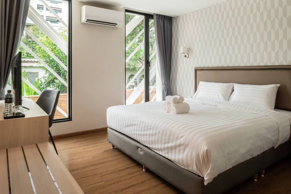 A modern hotel room with a large bed, a desk, and a view of greenery through the windows.