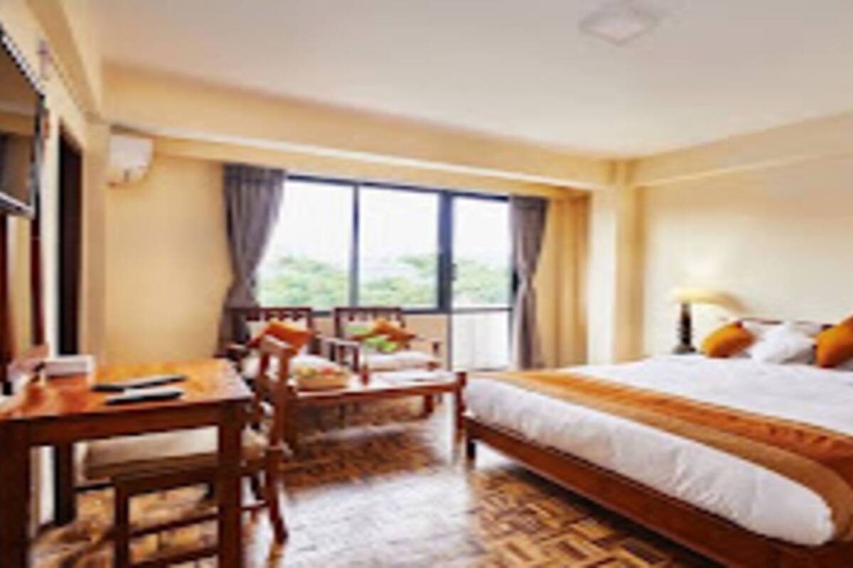 A hotel room with a large bed, a desk, and a chair. There is a window with curtains, and the room has a wooden floor.