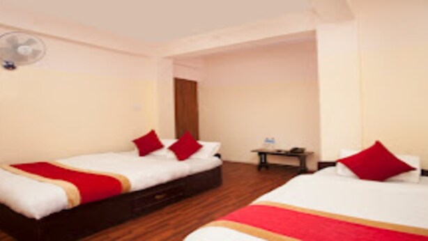 A hotel room with two beds, each with red and white bedding, a wooden floor, a small table with a lamp, and a wall-mounted fan. - MeroStay 122 Natraj Restaurant &Bar (Kathmandu)