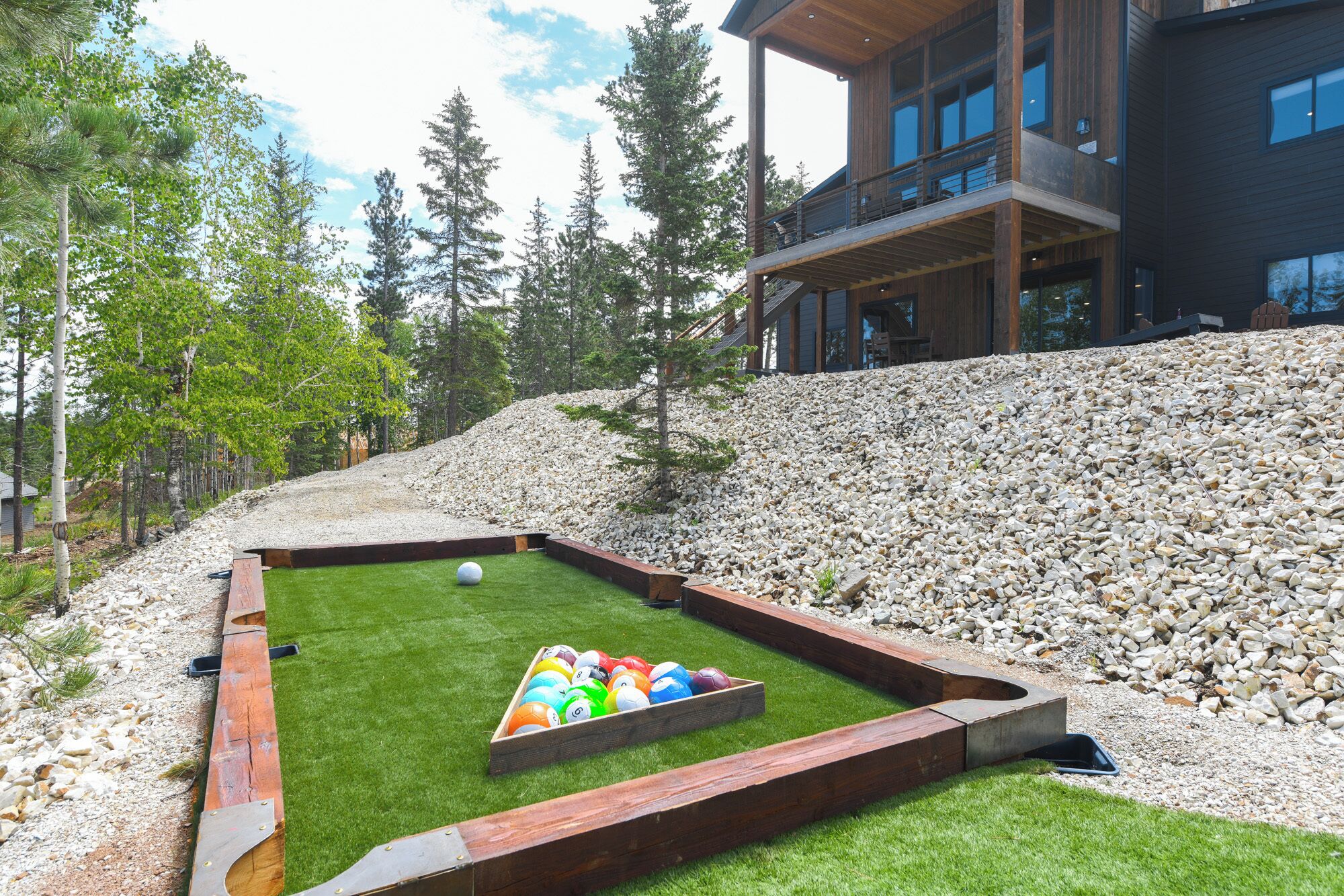 A pool table with a green felt surface and a wooden border, surrounded by a gravel area and trees.