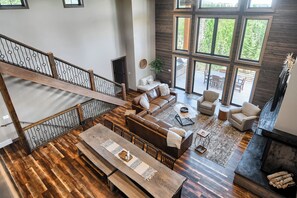 A spacious living area with a wooden staircase, a large window overlooking greenery, and a cozy seating arrangement. - Incredible 5000+ Sq. Ft. Lodge w/ Large Outdoor Space w/Hot Tub and Fire Pit! (Lead)