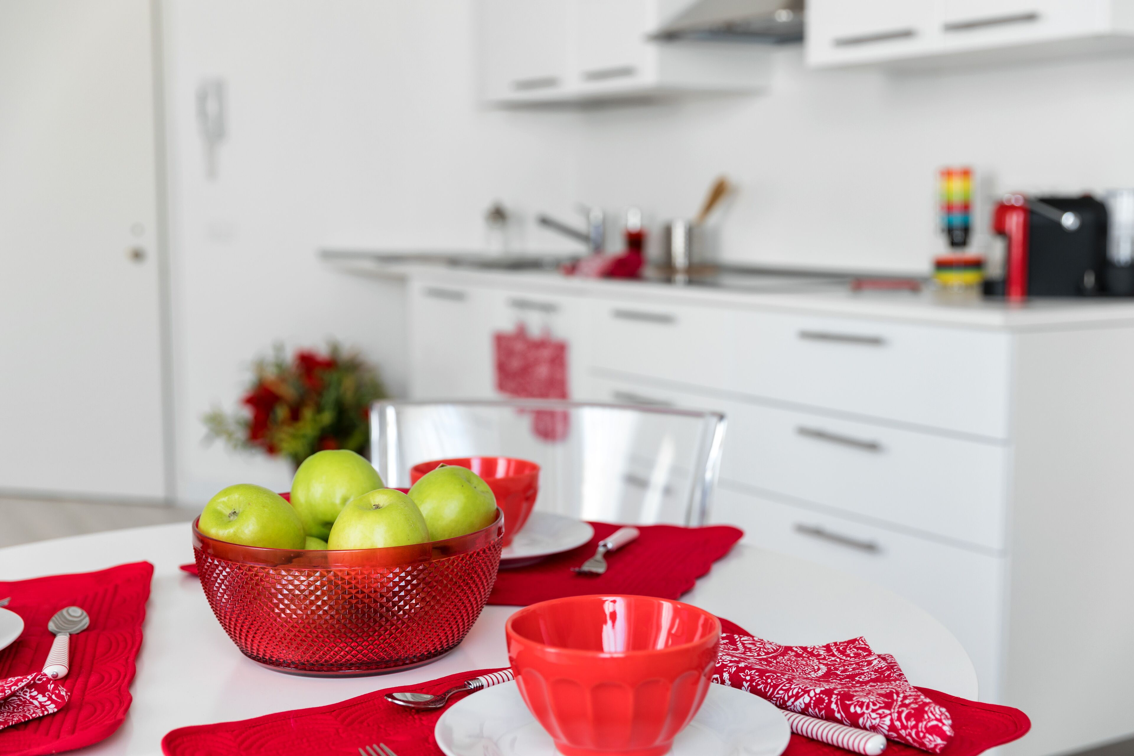Une cuisine avec des meubles blancs, un saladier rouge contenant des pommes et une tasse rouge posée sur la table.