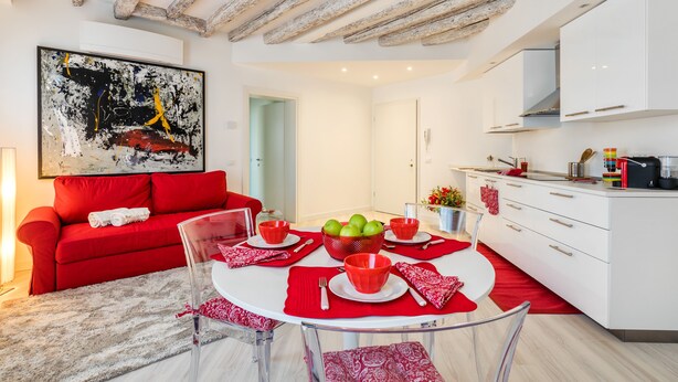 Un espacio habitable moderno con un sofá rojo, una mesa de comedor de cristal con individuales rojos y una cocina con armarios blancos.