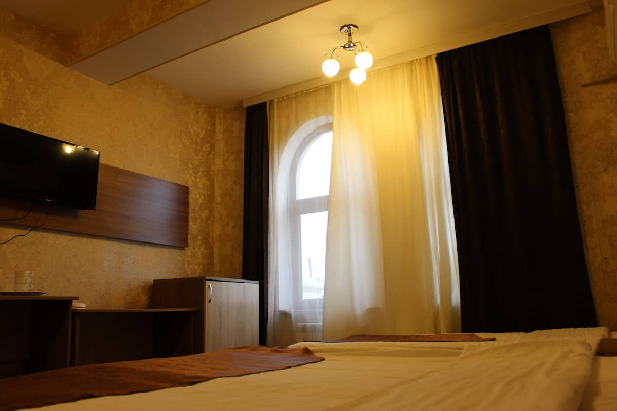 A hotel room with a bed, a TV mounted on the wall, a desk with a lamp, and a window with curtains.