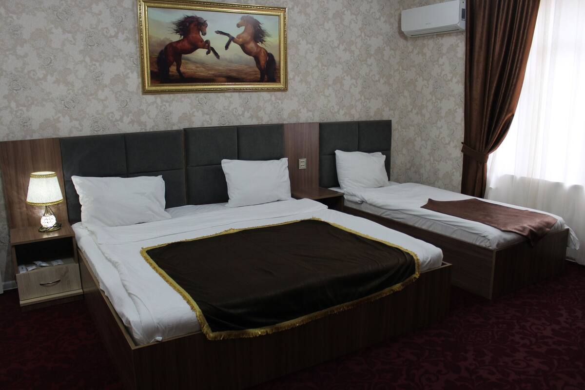 A hotel room with two beds, a nightstand with a lamp, a painting of horses, and a patterned carpet.