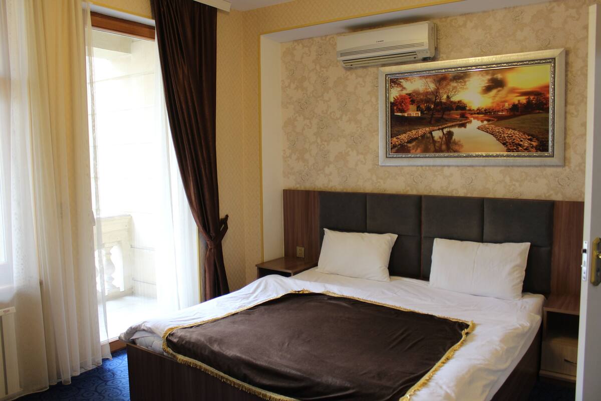A hotel room with a bed, a nightstand, a wall-mounted air conditioner, and a window with curtains.