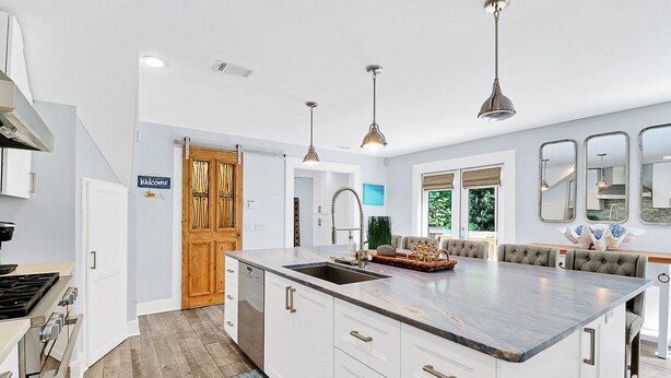 A modern kitchen with a large island, white cabinetry, and a built-in oven. There is a wooden door with a 'Welcome' sign, a dining area with a bench, and a large window with a view of trees.