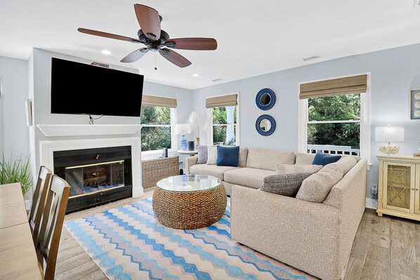 A living room with a fireplace, a ceiling fan, a television, and a large window with a view of trees.