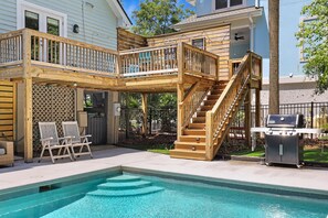 A backyard with a pool, wooden deck, outdoor seating, and a barbecue grill.
