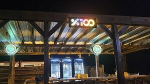 Uma área de estar ao ar livre rústica, com vigas de madeira e teto de metal, com um letreiro de neon e máquinas de venda automática iluminadas.