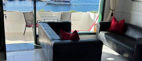 Un salón moderno con un sofá de cuero negro, un cojín rojo y vistas al mar y barcos a través de amplias ventanas.