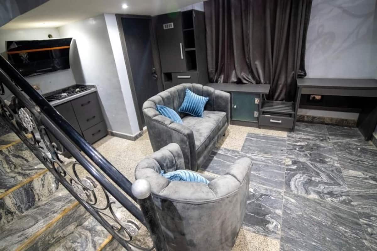 A modern living room with a grey sofa, a grey armchair, and a small ottoman. There is a staircase with a black railing and a flat-screen TV mounted on the wall.