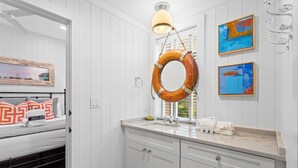 A bathroom with white paneled walls, a wooden-framed mirror, a round life preserver hanging above the sink, and two abstract paintings on the wall. - Goodnight Moon - Newly Renovated 30A Cottage in Historic Grayton Beach! (Grayton Beach)