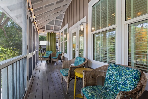 A covered porch with wicker furniture and a yellow table, large windows, and a view of greenery.
