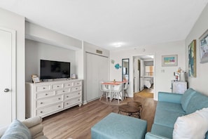 A living room with a TV, white dresser, teal sofa, and a dining area with a table and chairs.