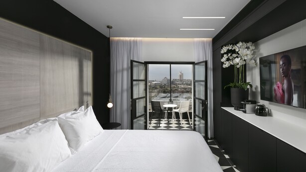 Una habitación de hotel moderna con una cama grande, un balcón con vista a la ciudad, una zona de comedor y un televisor en la pared.