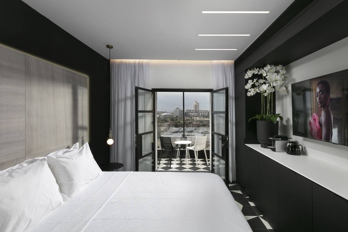 Une chambre d’hôtel moderne dotée d’un grand lit, d’un balcon offrant une vue sur la ville, d’un coin repas et d’un téléviseur mural.