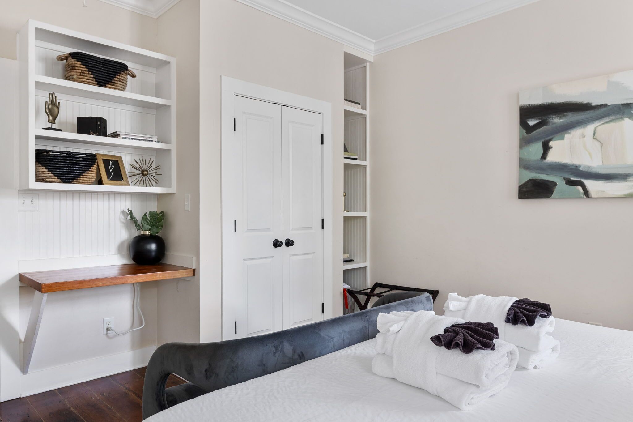 A hotel room with a bed, a wooden shelf with decorative items, a white wardrobe, and a painting on the wall.
