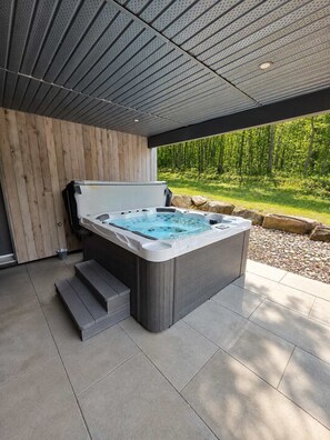 A modern outdoor hot tub with steps leading up to it, surrounded by a wooden wall and a covered area with a view of a bamboo forest.
