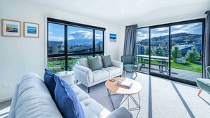 A modern living room with a sofa, armchairs, a coffee table, and large windows offering a view of a town and mountains. - Tranquil Heights Wanaka (Wānaka)