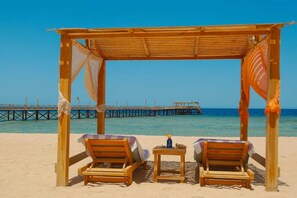A beach with a wooden gazebo, two lounge chairs, and a small table with a bottle.