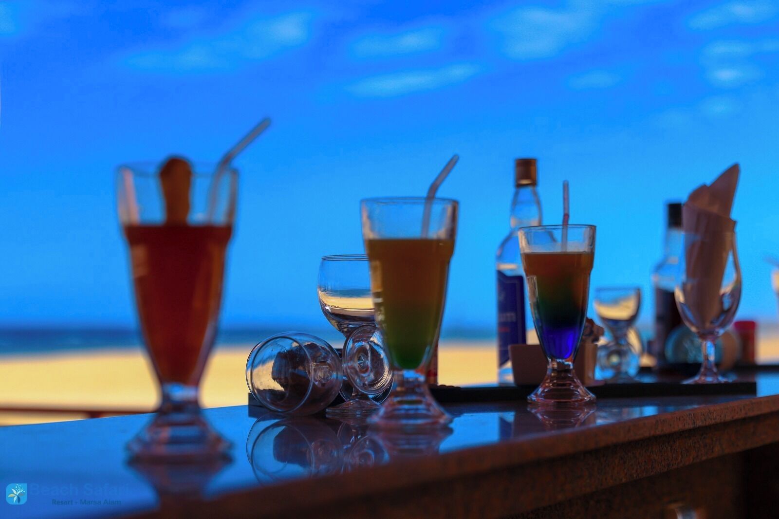 Un bar avec différents verres remplis de boissons et une bouteille posée sur une table, avec une vue sur la plage en arrière-plan.
