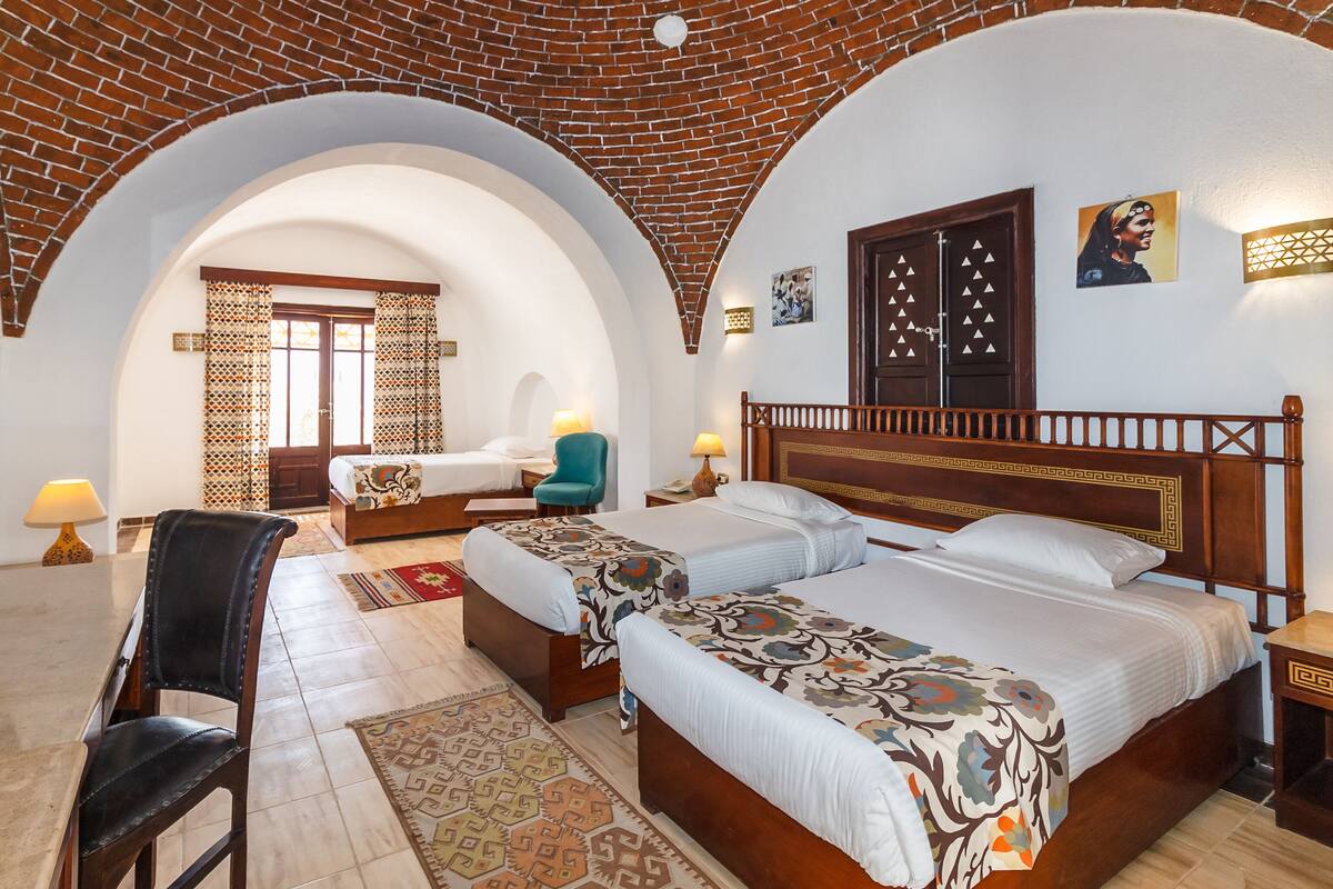 Ein Hotelzimmer mit zwei Betten, einem Schreibtisch und einem Stuhl. Die Decke besteht aus Ziegeln und die Tür aus Holz.
