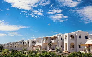 A row of white buildings with balconies and arched windows, surrounded by greenery and under a clear blue sky.