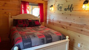 Un dormitorio de cabaña de madera acogedor, con cama, cortinas rojas y un cartel que dice 'Stay awhile' en la pared.