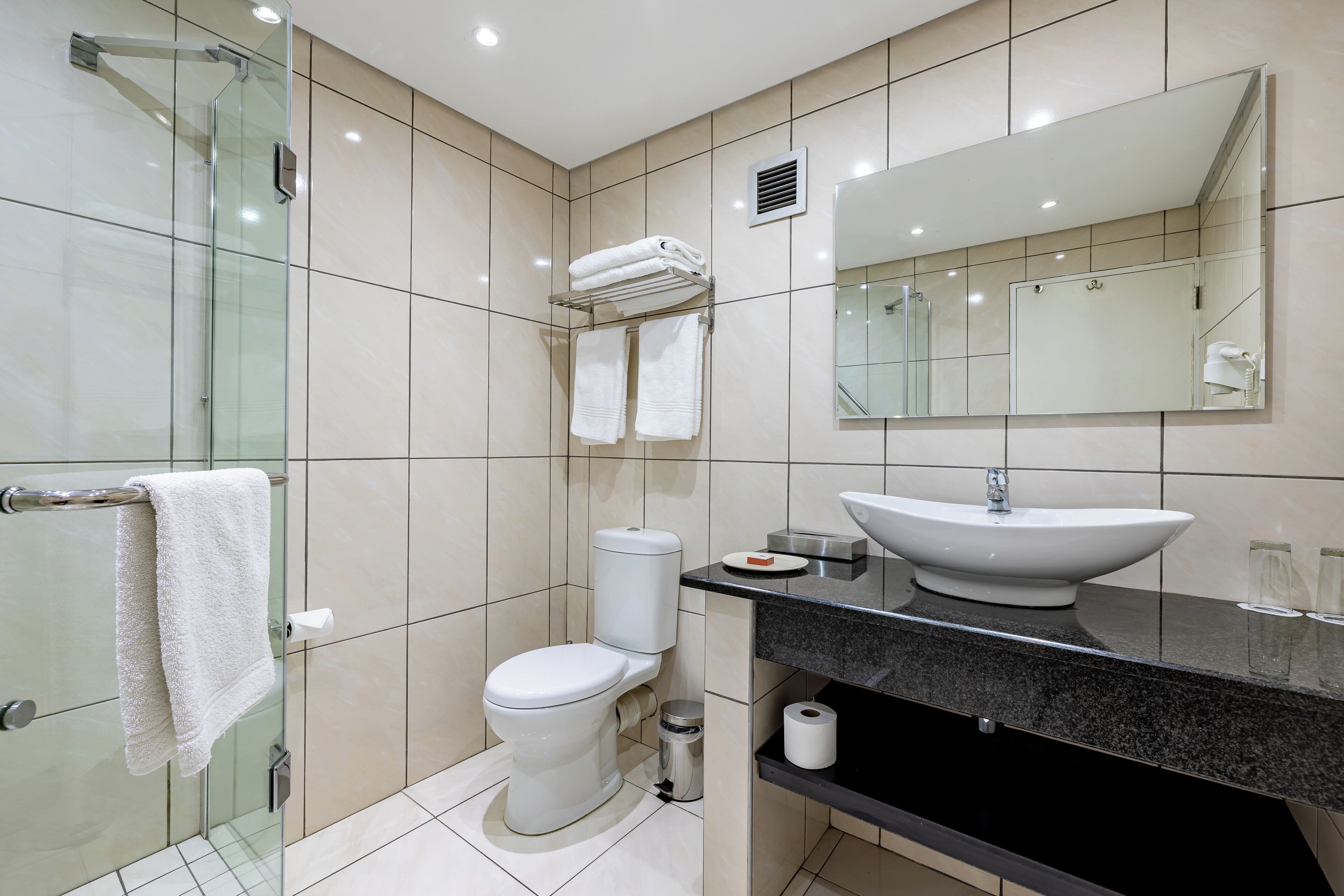 A modern bathroom with a glass shower enclosure, a toilet, a sink with a large mirror, and neatly folded towels on a shelf.