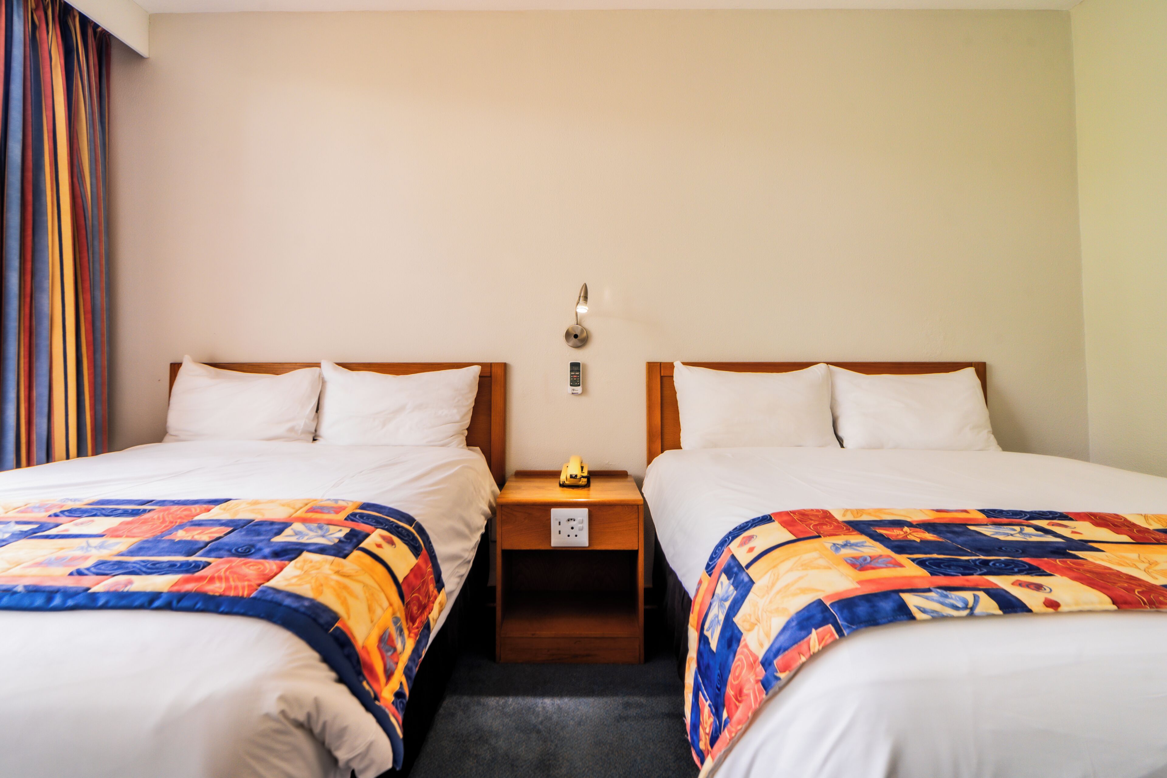 A hotel room with two beds, a nightstand with a phone, and a wall-mounted light.