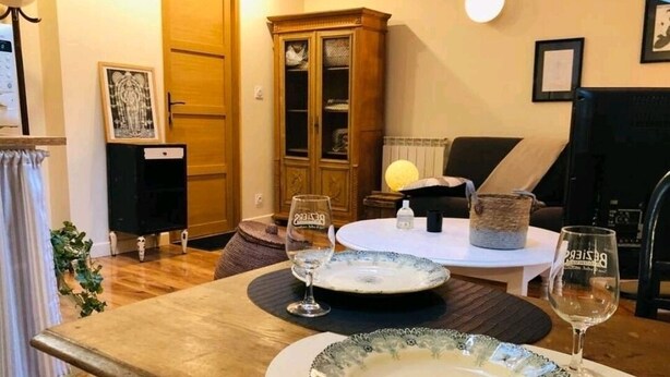 Une table à manger dressée avec des assiettes et des verres à vin, un buffet et une chaise, dans une pièce dotée d’un plancher en bois et où l’on aperçoit une porte au fond.