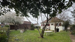Un jardín con una mesa blanca, un cobertizo de madera y una casa con contraventanas rojas.