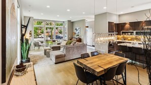 A modern kitchen with a wooden dining table and bar stools, a living area with a sofa and chairs, and a large window offering a view of the street.