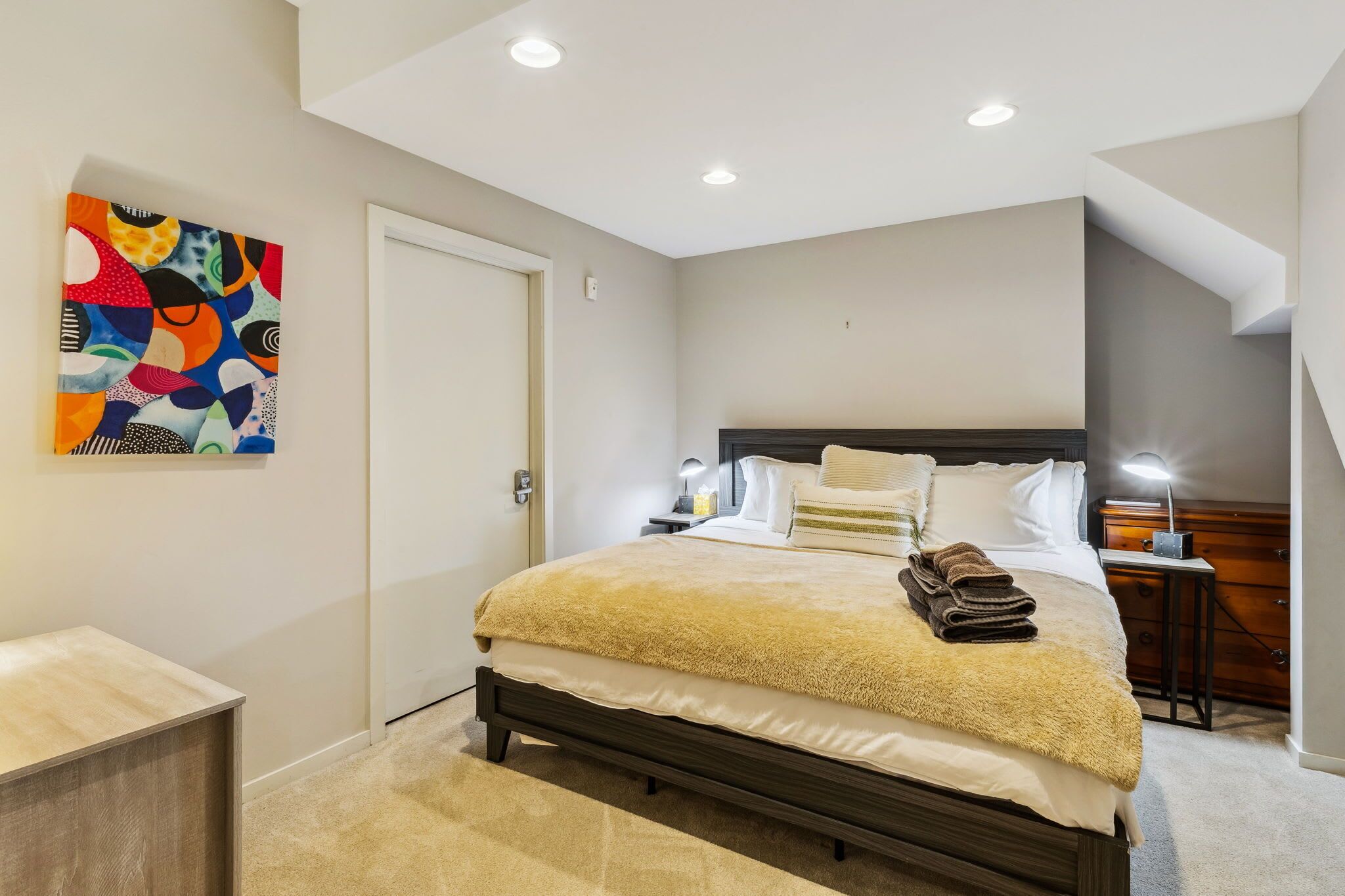 Un dormitorio moderno con una cama grande, una mesita de noche con lámpara, una mesita auxiliar de madera y un cuadro abstracto colorido en la pared.