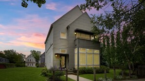 A modern two-story house with a large glass window on the second floor, surrounded by a well-maintained lawn and a wooden fence.