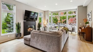 A spacious living room with a large grey sofa, a flat-screen TV mounted above a fireplace, and a piano in the corner.